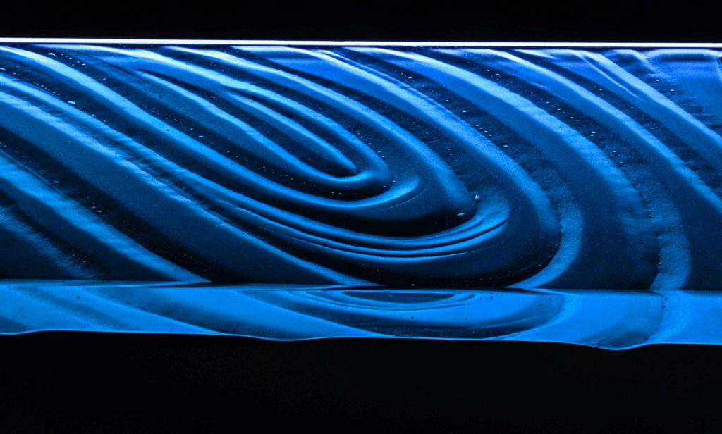 Close-up macro photograph of a hand-polished glass bar top edge with COB LED lighting in deep blue, showing lens-quality edge clarity and light refraction — Downing Designs Tampa