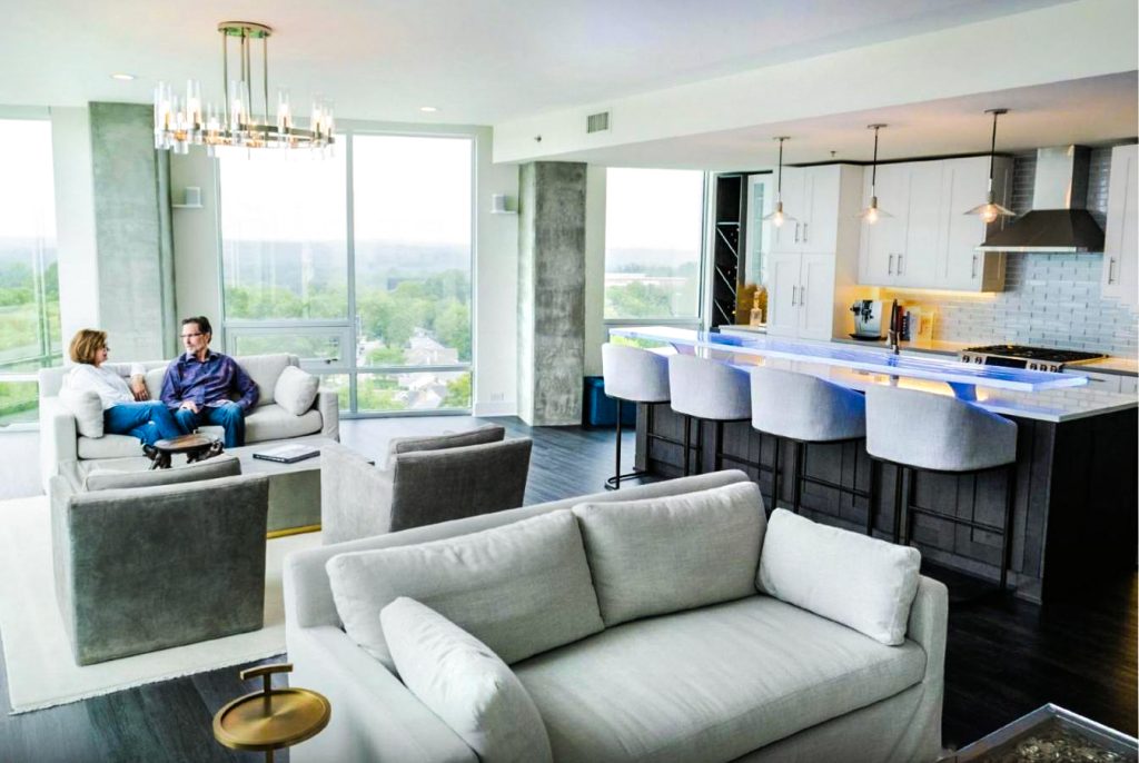Wide view of ninth-floor luxury condominium at Greenbridge Condominiums, Chapel Hill NC — custom LED-illuminated kiln-formed glass bar top by Downing Designs glowing blue in the kitchen, floor-to-ceiling city views beyond